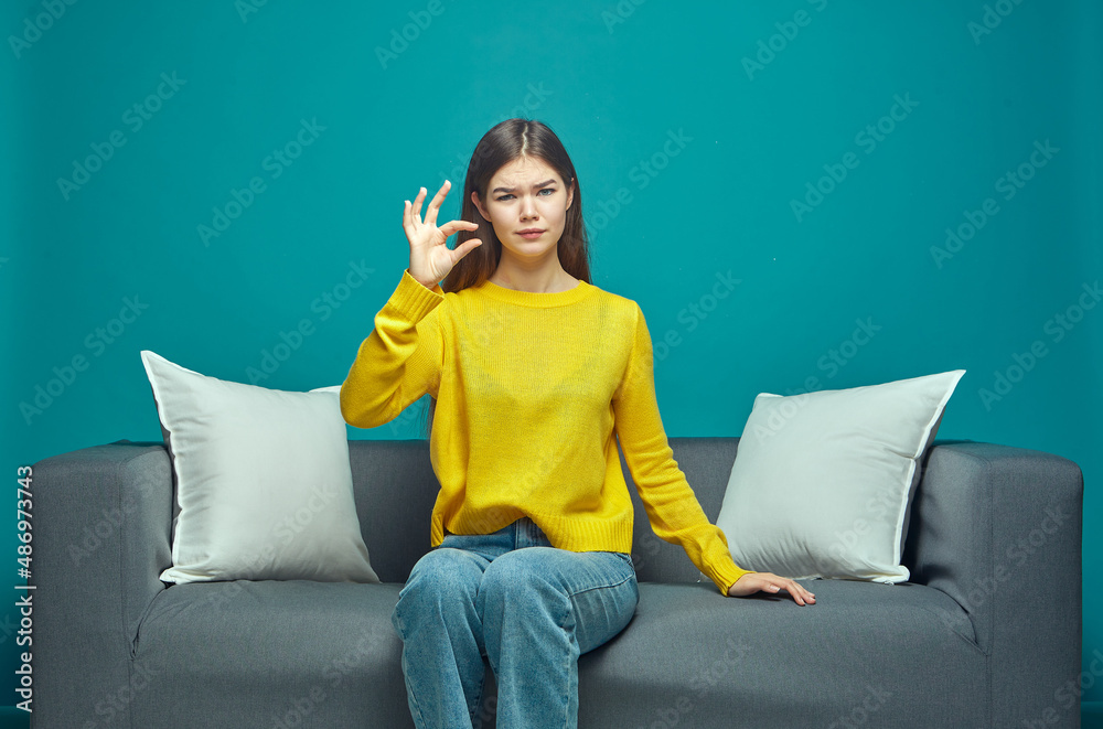 Displeased young girl showing little bit gesture, small size, dissatisfied with low rating, sitting on couch Displeased young girl showing little bit gesture, small size, dissatisfied with low rating, sitting on couch
