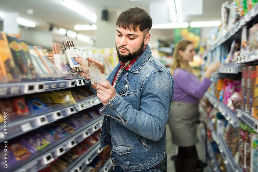 Bearded guy choosing chocolate on shelves in grocery store