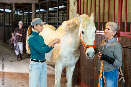 Senior and younger caucasian women braiding mane of white horse in barn.