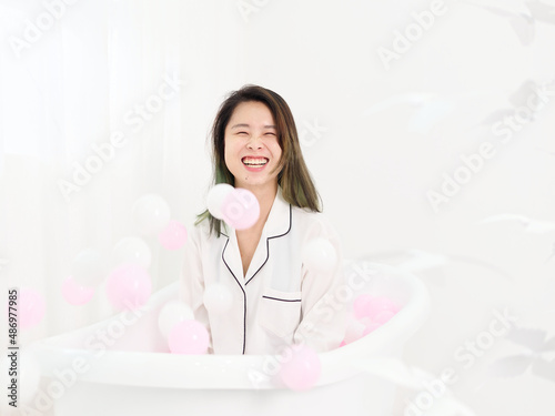 Portrait of young Asian woman sitting in bathtub and smiling at flying ocean ball in air, beautiful Chinese girl with green hairs in white shirt enjoy her happy time carefreely, half length shot. photo