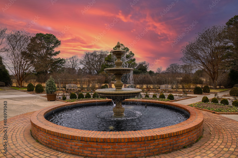 a circular water fountain in the garden surrounded by red brick, bare winter trees and lush green trees with red sky and powerful clouds at sunset at Memphis Botanic Garden in Memphis Tennessee USA - obrazy, fototapety, plakaty 