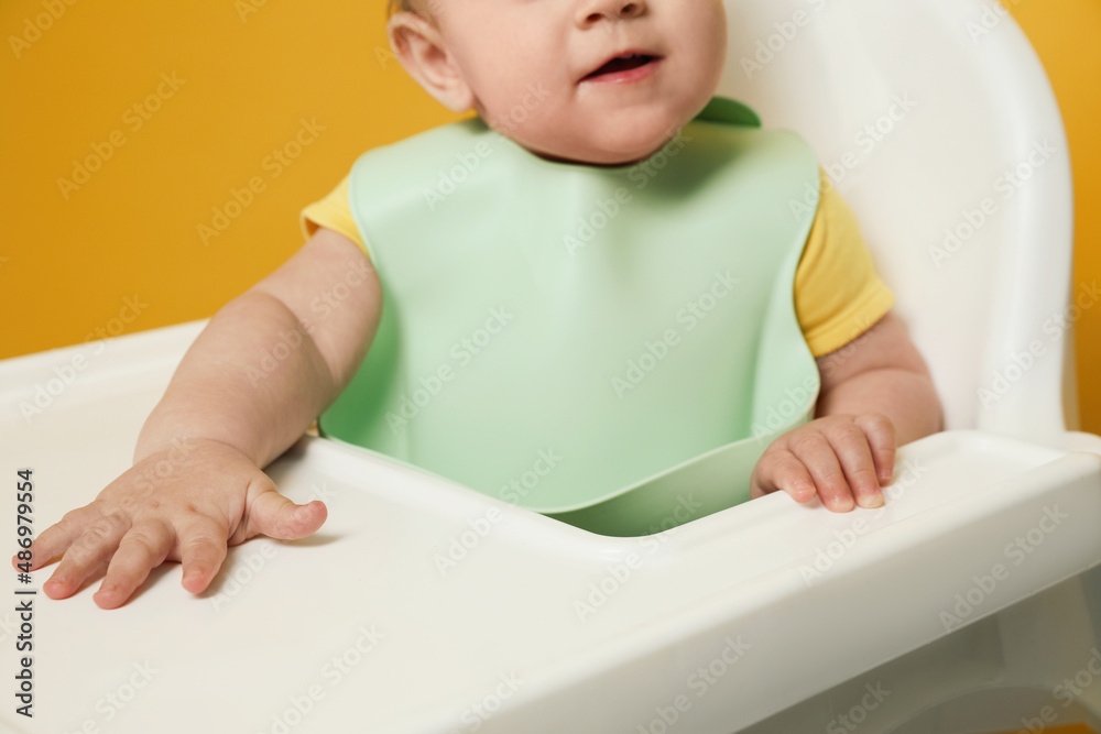 Cute little baby wearing bib in highchair on yellow background, closeup Cute little baby wearing bib in highchair on yellow background, closeup