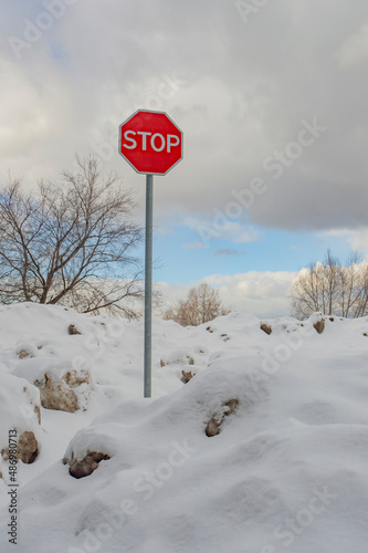 Stop road sign on a road blocked with huge snowdrifts. Blue winter sky with grey clouds, naked trees © Dmitry