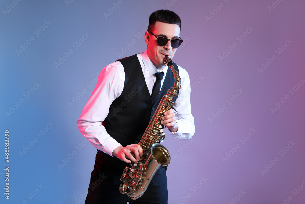 Fototapeta premium Young man in elegant outfit playing saxophone on color background