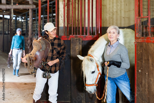 Positive Asian horsewoman holding saddle while elderly female stable keeper leading white horse out of stall by bridle..
