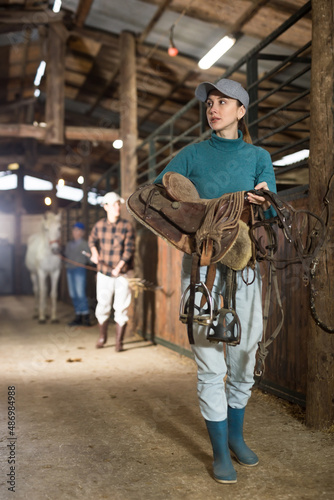 Portrait of young horsewoman walking in stable with saddle in hands, about to harness horse for riding