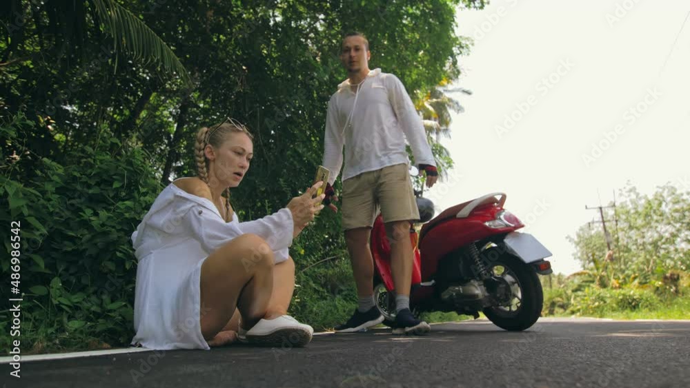 Woman on red motorbike in white clothes drive on forest road trail trip. One girl caucasian tourist sit, relax, rest, near scooter. Use a smartphone, take a selfie. Motorcycle rent. Woman on red motorbike in white clothes drive on forest road trail trip. One girl caucasian tourist sit, relax, rest, near scooter. Use a smartphone, take a selfie. Motorcycle rent.