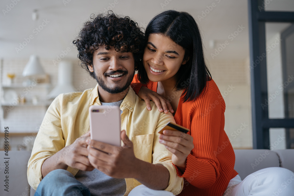 Smiling multiracial couple holding credit card shopping online, ordering food looking at digital screen sitting at cozy home. Happy freelancers receive payment, successful business 