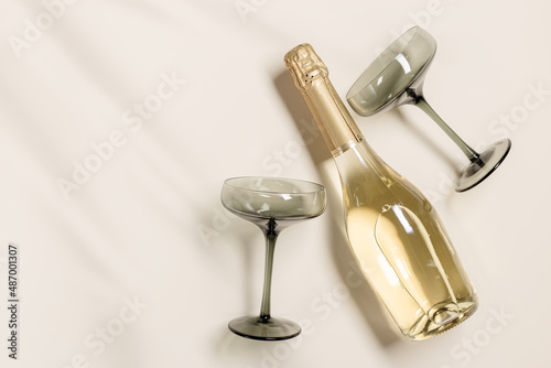 Champagne glasses tinted grey glass, Champagne or sparkling wine bottle on beige background. Festive drink concept, glare and sunlight. Modern wine colored glasses. Creative flat lay
