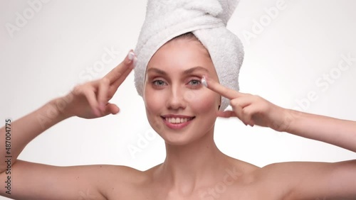 Funny attractive woman apply skincare cream on face look at the camera, healthy young lady wrap towel on head put moisturizing lifting facial creme, healthy skin care treatment concept.