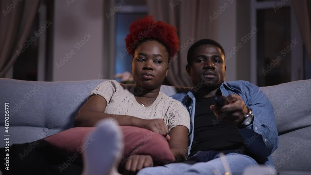 Young happy couple turning on tv and sitting on couch in apartment interior spbas. Close-up view of african american people watch movie and hug, sit on sofa in dark living room, handsome guy holds Young happy couple turning on tv and sitting on couch in apartment interior spbas. Close-up view of african american people watch movie and hug, sit on sofa in dark living room, handsome guy holds