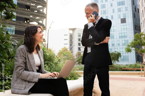 Young caucasian woman and adult man work outside office buildings using laptop and mobile phone.