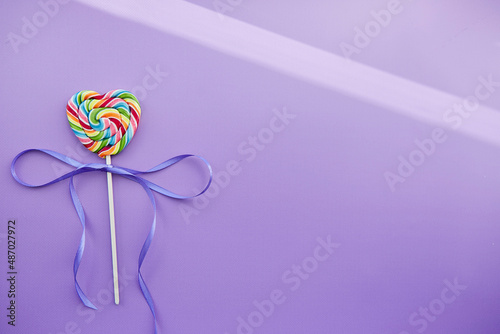 Rainbow heart shaped sweet lollipop on trendy violet background with festive ribbon and shadows. Copy space. Minimalist present concept.