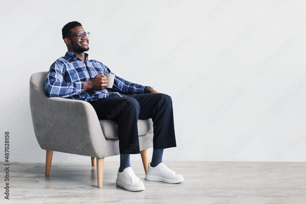 Coffee break. Dreamy young African American man sitting in armchair, enjoying cup of hot beverage against white wall Coffee break. Dreamy young African American man sitting in armchair, enjoying cup of hot beverage against white wall