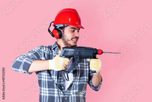 Young worker with a drill and helmet, on pink background.