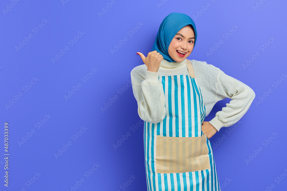 Portrait of smiling young housewife woman in hijab and apron looking at camera, pointing aside with finger isolated on purple background. People housewife muslim lifestyle concept Portrait of smiling young housewife woman in hijab and apron looking at camera, pointing aside with finger isolated on purple background. People housewife muslim lifestyle concept