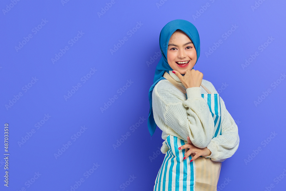 Portrait of smiling young housewife woman in hijab and apron looking at camera, pointing aside with finger isolated on purple background. People housewife muslim lifestyle concept Portrait of smiling young housewife woman in hijab and apron looking at camera, pointing aside with finger isolated on purple background. People housewife muslim lifestyle concept