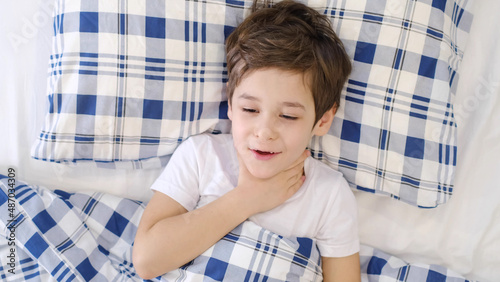 Sick boy touching his painful neck, sore throat and flu, throat infection. toddler boy is lying in bed and blowing nose into tissue paper at home. Allergy, flu