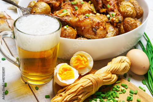 lunch in rural style, tasty food - glass of beer, fried chicken meat and potatoes, garlic and green onions, cheese, farm eggs and salt, cooked food on a white wooden boards