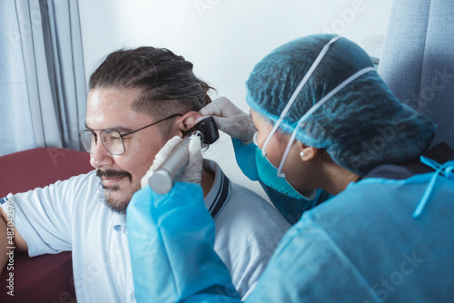 A doctor in full PPE gear checks the ear drum of a male patient with an otoscope, also known as an auriscope. Otoscopy procedure at the emergency ward of a hospital.