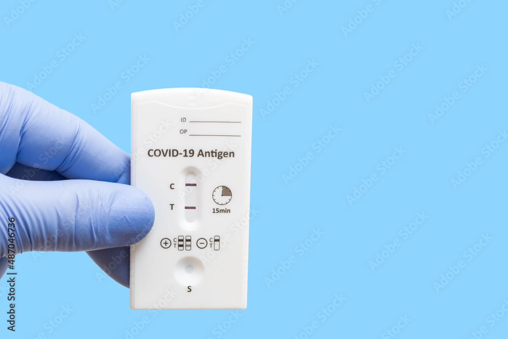 Hand in a medical glove holds rapid antigen test kit for viral disease COVID-19 with positive result, blue background with copy space