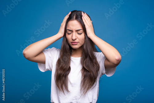 Attractive caucasian or arab brunette girl in a white t-shirt holding her hands behind her head because of a headache isolated on a blue studio background.