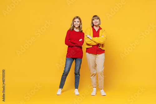 Full body smiling happy cool woman 50s in red shirt have fun with teenager girl 12-13 years old. Grandmother granddaughter hold hands crosed folded isolated on plain yellow background. Family concept