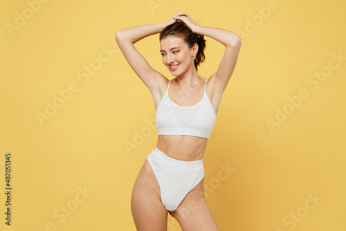 Smiling minded attractive young brunette woman 20s wearing white underwear with perfect fit body standing ties hair in ponytail look camera isolated on plain yellow color background studio portrait