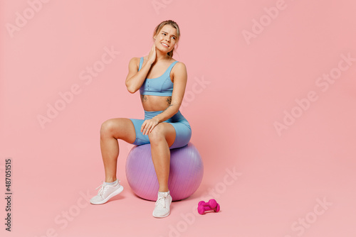 Full length young sporty fitness trainer instructor woman wear blue tracksuit spend time in home gym sit on fitball hold neck suffer from pain isolated on plain pink background. Workout sport concept.