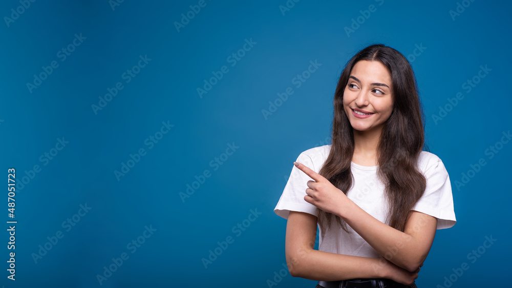 Attractive caucasian or arab brunette girl in white t-shirt pointing with finger at copy space for advertising isolated on blue studio background. Attractive caucasian or arab brunette girl in white t-shirt pointing with finger at copy space for advertising isolated on blue studio background.