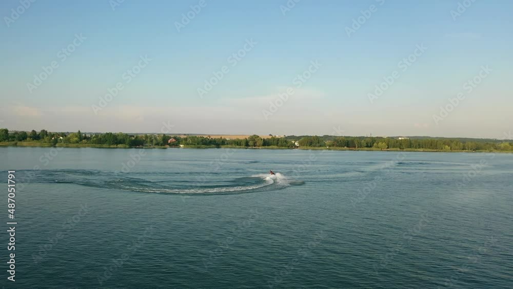 Aerial view from drone on active man in life vest doing various tricks while riding jet ski on local lake. Summer activity on water of sporty person.