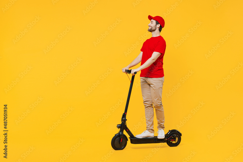 Full size side view delivery guy employee man in red cap T-shirt uniform workwear work as dealer courier ride electric kick scooter isolated on plain yellow background studio portrait Service concept Full size side view delivery guy employee man in red cap T-shirt uniform workwear work as dealer courier ride electric kick scooter isolated on plain yellow background studio portrait Service concept