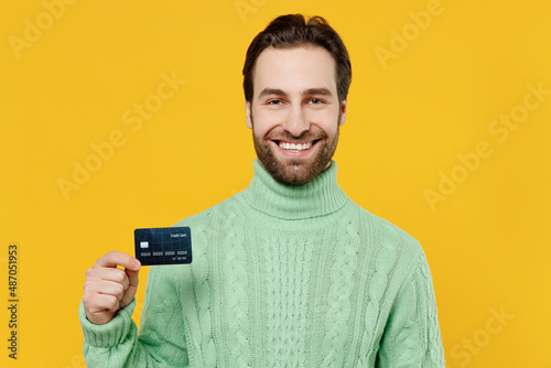 Young cheerful satisfied cool rich smiling happy man 20s wear mint knitted sweater hold in hand credit bank card isolated on plain yellow background studio portrait. People lifestyle fashion concept.