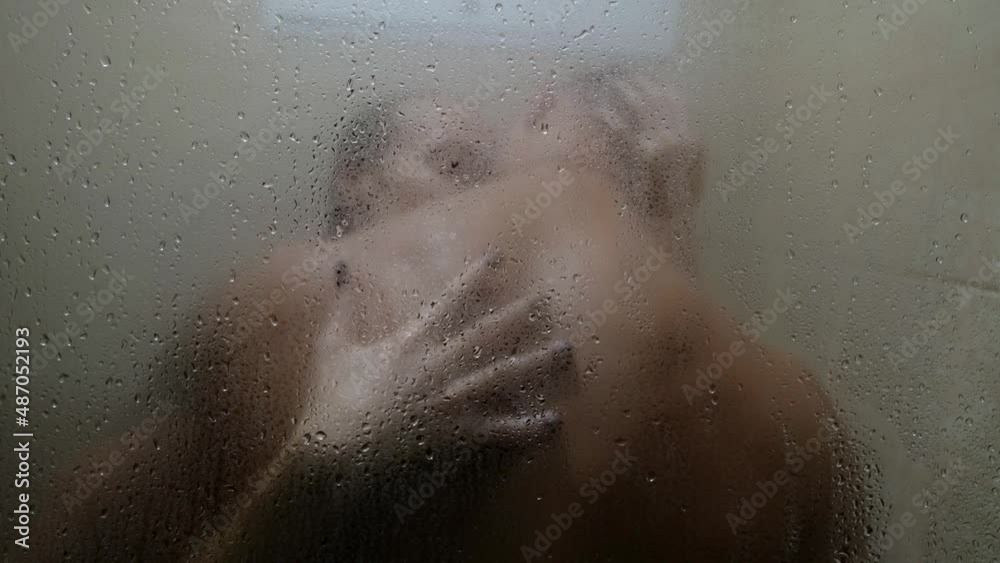 the couple is kissing in the shower and is seen out of focus through the glass with drops. women's hands slide down the man's back the couple is kissing in the shower and is seen out of focus through the glass with drops. women's hands slide down the man's back