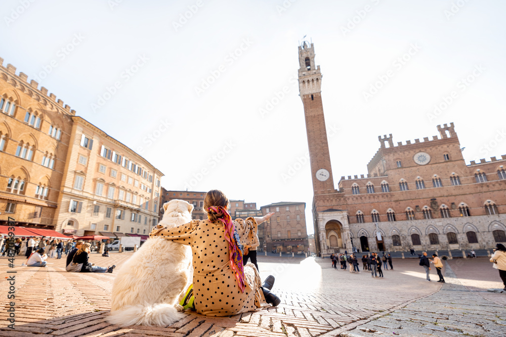 Naklejka premium Woman sitting with her dog on main square of Siena city with a town hall on background. View from the backside. Concept of travel on tuscany and friendship with pet