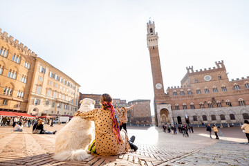 Woman sitting with her dog on main square of Siena city with a town hall on background. View from the backside. Concept of travel on tuscany and friendship with pet