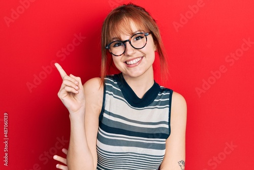 Fototapeta Naklejka Na Ścianę i Meble -  Redhead young woman wearing casual clothes and glasses smiling happy pointing with hand and finger to the side