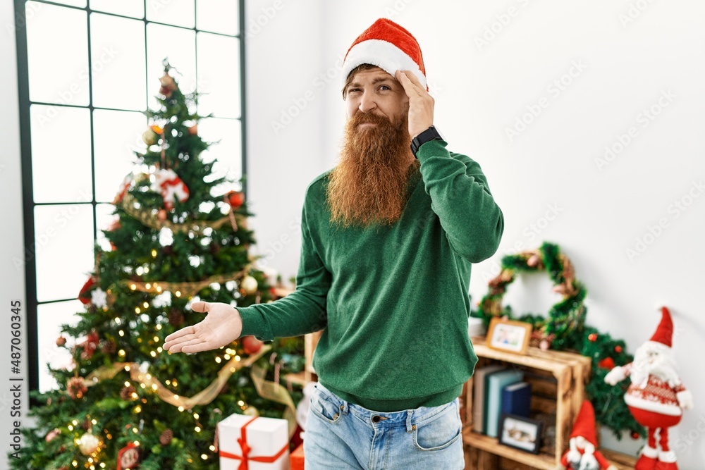 Redhead man with long beard wearing christmas hat by christmas tree confused and annoyed with open palm showing copy space and pointing finger to forehead. think about it.