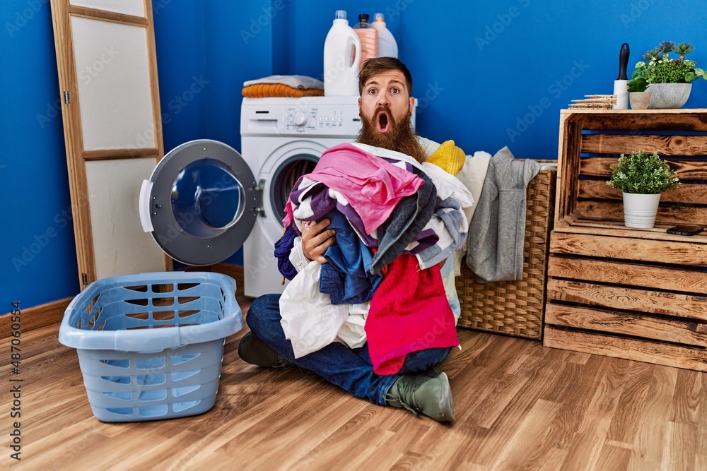 Redhead man with long beard putting dirty laundry into washing machine afraid and shocked with surprise and amazed expression, fear and excited face. Redhead man with long beard putting dirty laundry into washing machine afraid and shocked with surprise and amazed expression, fear and excited face.