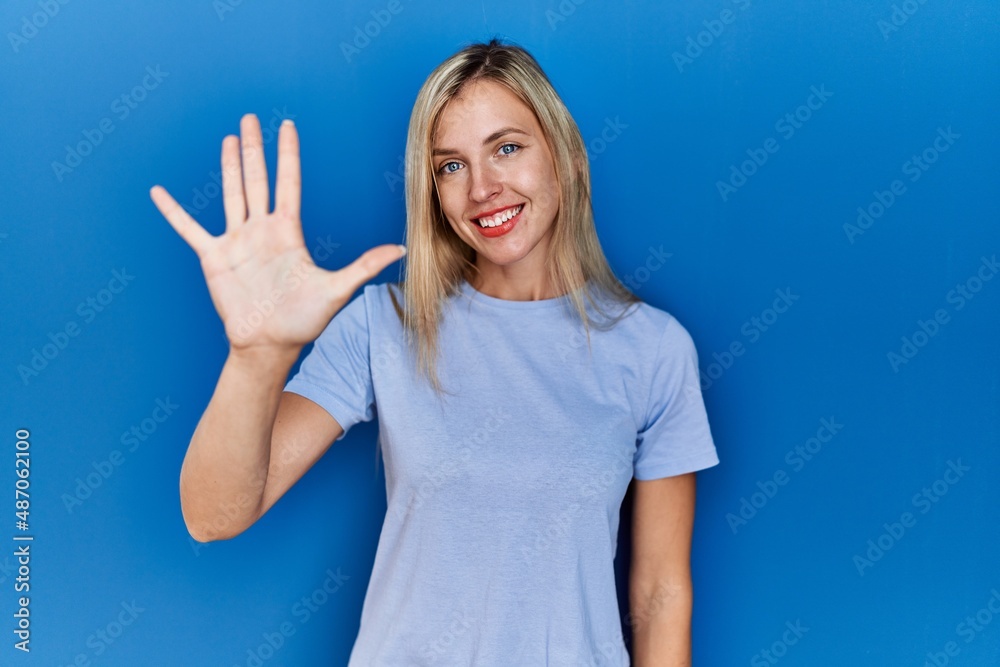 Beautiful blonde woman wearing casual t shirt over blue background showing and pointing up with fingers number five while smiling confident and happy. Beautiful blonde woman wearing casual t shirt over blue background showing and pointing up with fingers number five while smiling confident and happy.