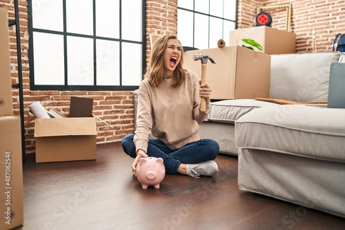 Young woman holding piggy bank and hammer moving to a new home angry and mad screaming frustrated and furious, shouting with anger. rage and aggressive concept.