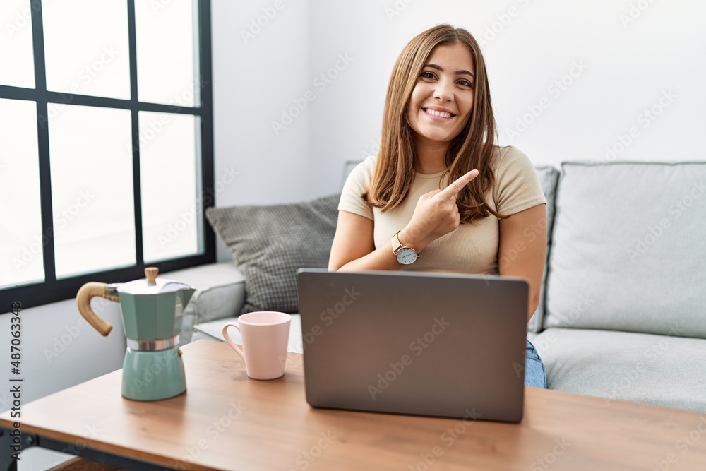 Young brunette woman using laptop at home drinking a cup of coffee cheerful with a smile of face pointing with hand and finger up to the side with happy and natural expression on face Young brunette woman using laptop at home drinking a cup of coffee cheerful with a smile of face pointing with hand and finger up to the side with happy and natural expression on face