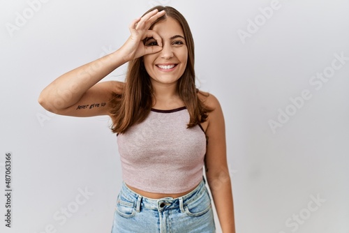 Young brunette woman standing over isolated background doing ok gesture with hand smiling, eye looking through fingers with happy face. © Krakenimages.com