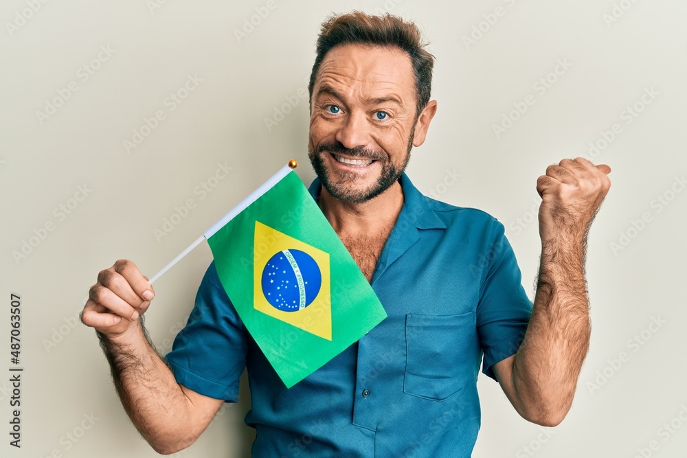 Middle age man holding brazil flag pointing thumb up to the side smiling happy with open mouth