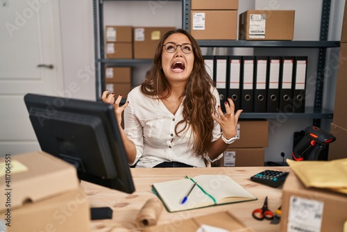 Young hispanic woman working at small business ecommerce crazy and mad shouting and yelling with aggressive expression and arms raised. frustration concept.