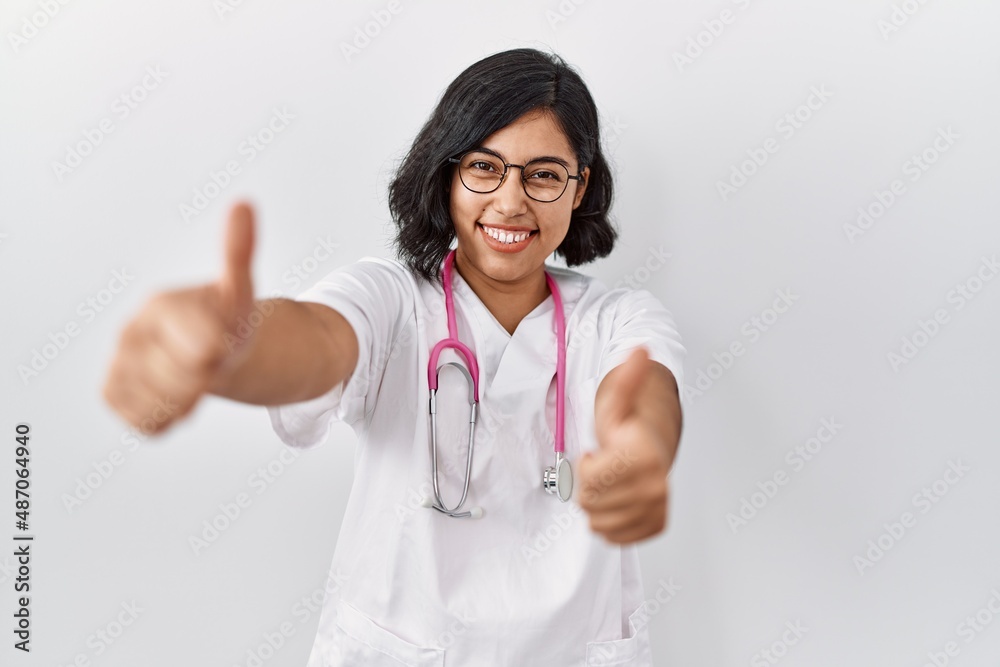 Young hispanic doctor woman wearing stethoscope over isolated background approving doing positive gesture with hand, thumbs up smiling and happy for success. winner gesture. Young hispanic doctor woman wearing stethoscope over isolated background approving doing positive gesture with hand, thumbs up smiling and happy for success. winner gesture.