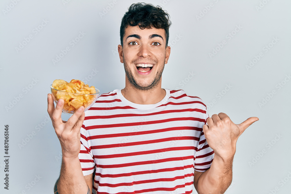 Young hispanic man holding potato chip pointing thumb up to the side smiling happy with open mouth Young hispanic man holding potato chip pointing thumb up to the side smiling happy with open mouth