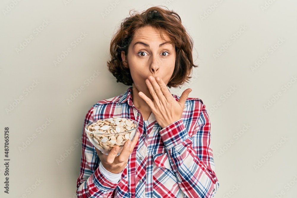 Young brunette woman holding bowl with pumpkin seeds covering mouth with hand, shocked and afraid for mistake. surprised expression Young brunette woman holding bowl with pumpkin seeds covering mouth with hand, shocked and afraid for mistake. surprised expression
