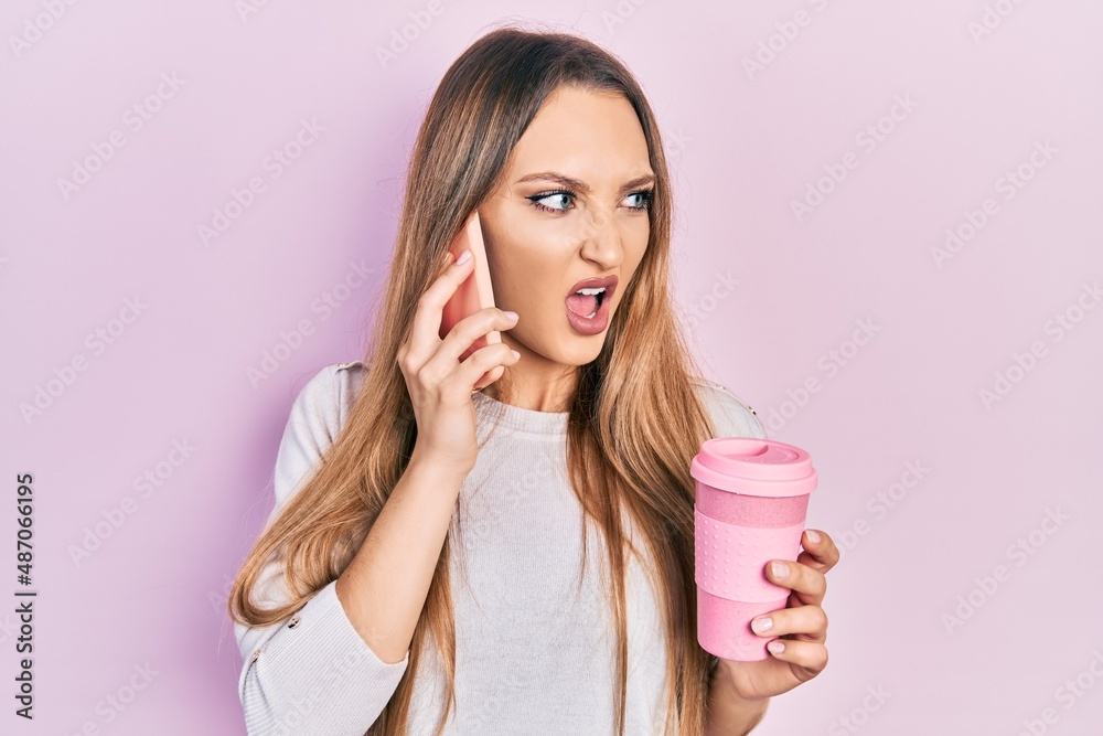 Young blonde girl using smartphone and drinking a cup of coffee angry and mad screaming frustrated and furious, shouting with anger. rage and aggressive concept. Young blonde girl using smartphone and drinking a cup of coffee angry and mad screaming frustrated and furious, shouting with anger. rage and aggressive concept.