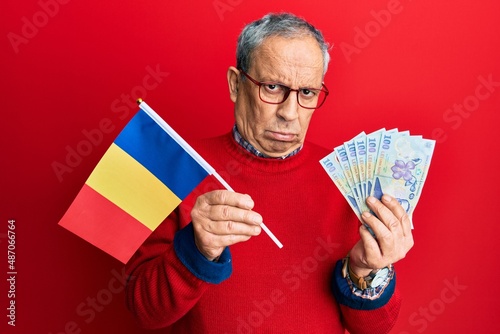 Handsome senior man with grey hair holding romania flag and leu banknotes skeptic and nervous, frowning upset because of problem. negative person.